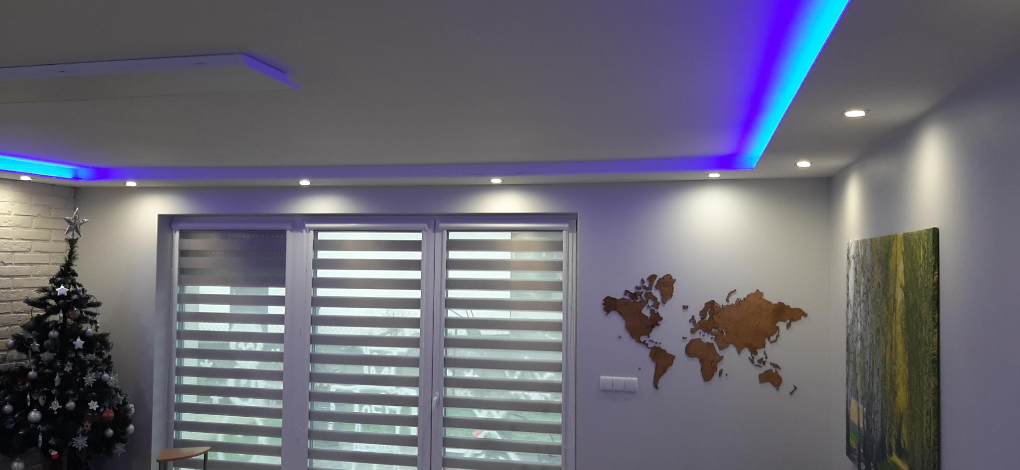 Wykończenie wnętrza w Oławie z podwieszanym sufitem z niebieskim oświetleniem LED, drewnianą mapą świata na ścianie i zasłoniętym oknem.