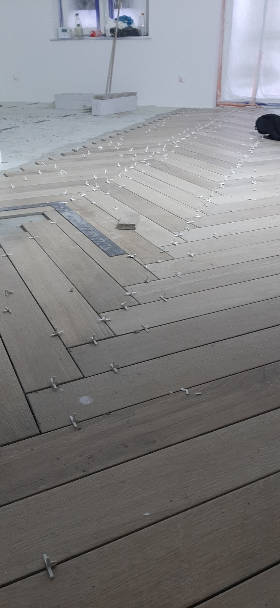 Ułożona w jodełkę drewniana podłoga z dystansami krzyżykowymi, widoczny fragment ściany i okno zabezpieczone folią malarską.