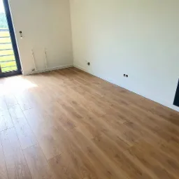 Jasne panele podłogowe ułożone w pokoju z oknem balkonowym, ściany pomalowane na biało i jasnozielono, z fragmentem ściany pomalowanym na czarno, widoczna skrzynka narzędziowa przy ścianie.