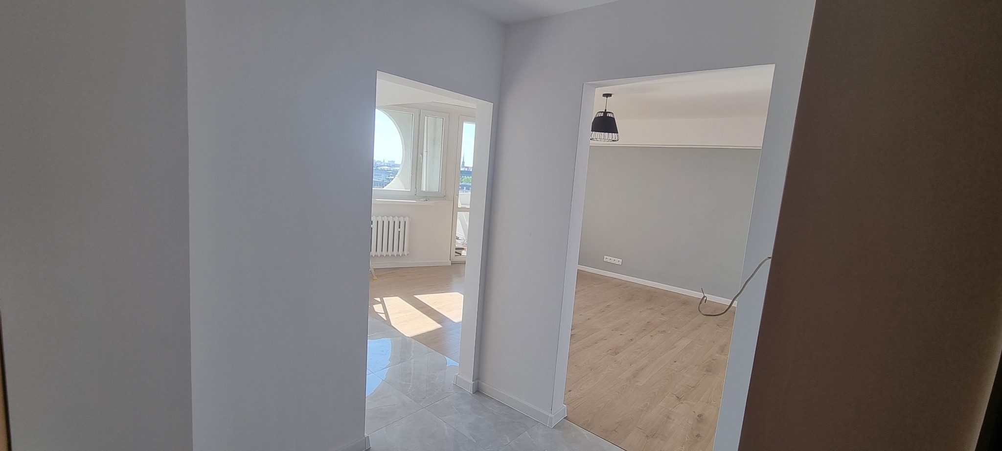 Widok z korytarza na świeżo pomalowane, jasnoszare ściany i ułożoną podłogę w mieszkaniu, z widokiem na balkon z okrągłym oknem i grzejnikiem.