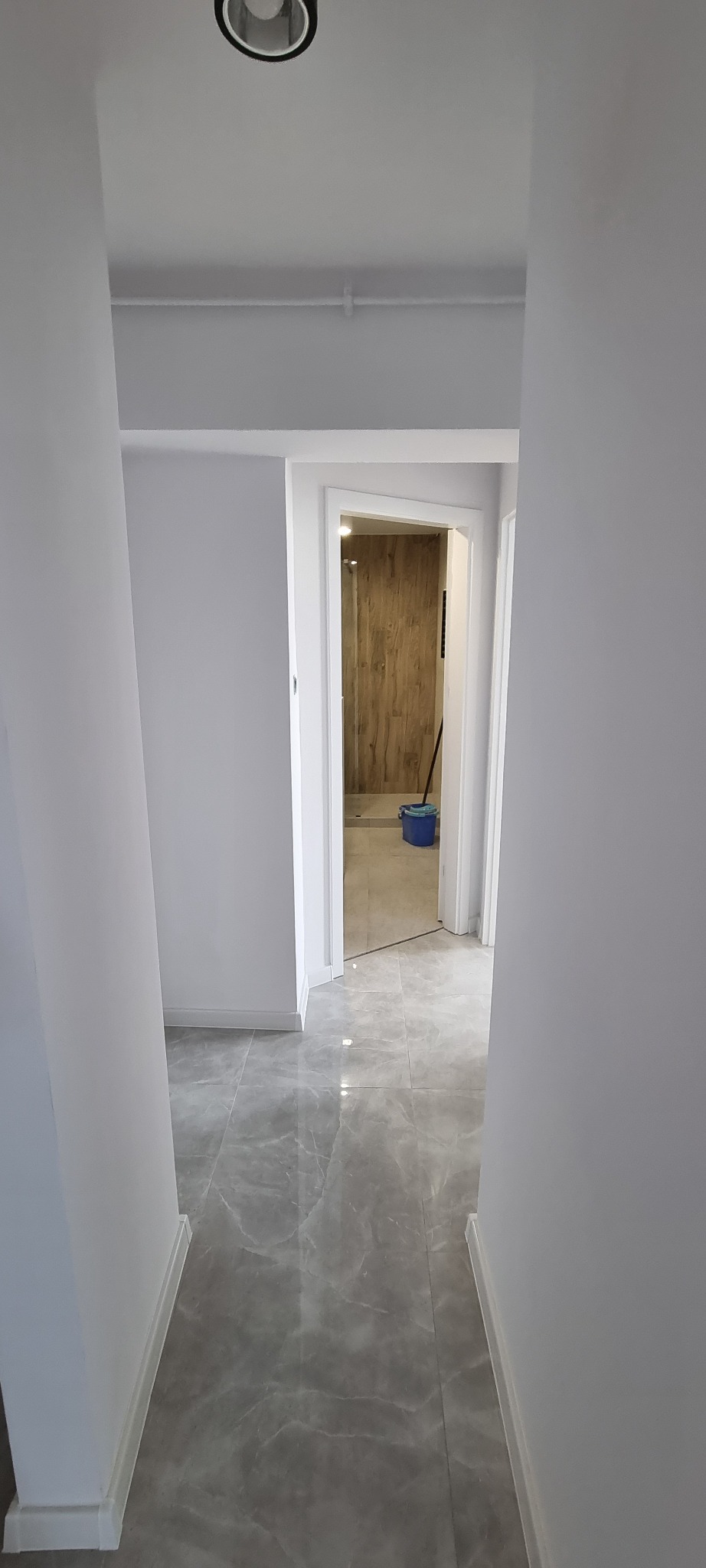 Korytarz po remoncie z szarymi płytkami podłogowymi imitującymi marmur, prowadzący do łazienki z drewnianą okładziną ścienną i niebieskim wiadrem.