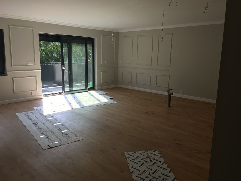 Jasne wnętrze z ułożoną drewnianą podłogą zabezpieczoną kartonem, dekoracyjne panele ścienne, przesuwne drzwi balkonowe wpadające światło słoneczne, wystające kable z sufitu.