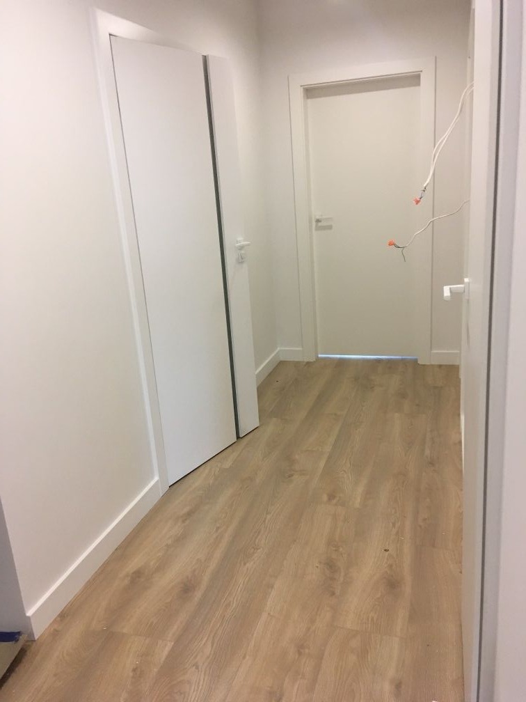 Wąski korytarz z panelami podłogowymi w odcieniach jasnego drewna, białe ściany i drzwi, widoczne kable wystające ze ściany przy jednym z wejść.