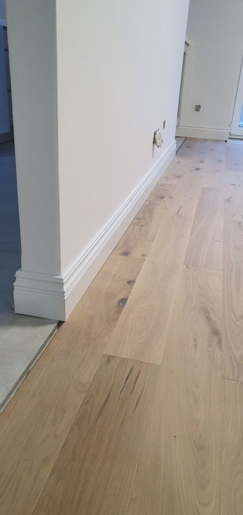 Jasny parkiet ułożony wzdłuż ściany z białą listwą przypodłogową, widoczne ślady po montażu gniazdka elektrycznego.