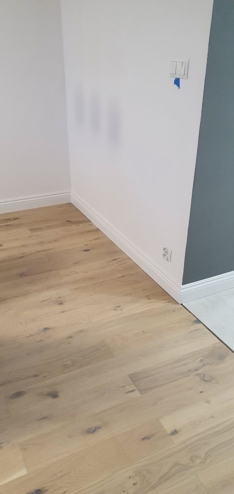 Jasny parkiet ułożony w pokoju z białymi ścianami i szarą ścianą akcentującą, widoczne listwy przypodłogowe i gniazdko elektryczne