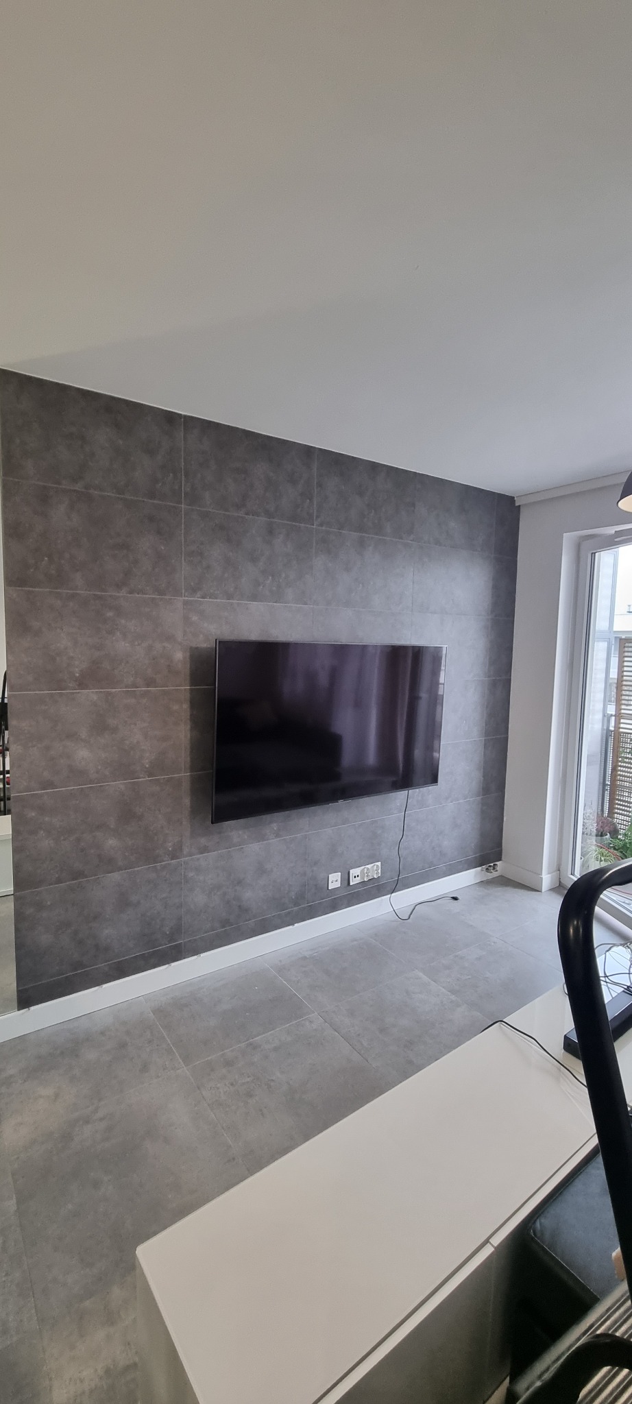 Salon z szarą ścianą z płytek imitujących beton, na której wisi czarny telewizor; widoczne gniazdka elektryczne i listwa przypodłogowa