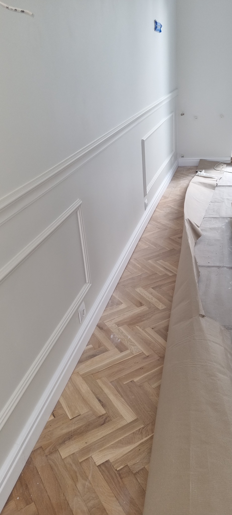 Ułożony parkiet w jodełkę, zabezpieczony częściowo papierem ochronnym, białe ściany z dekoracyjnymi listwami przypodłogowymi i gzymsami.