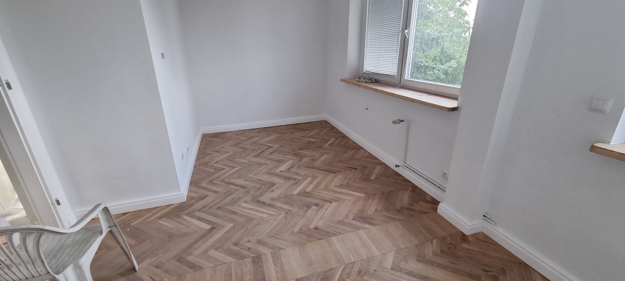 Wnętrze pokoju po remoncie z ułożonym parkietem w jodełkę, białe ściany, drewniany parapet i widok na zieleń za oknem.