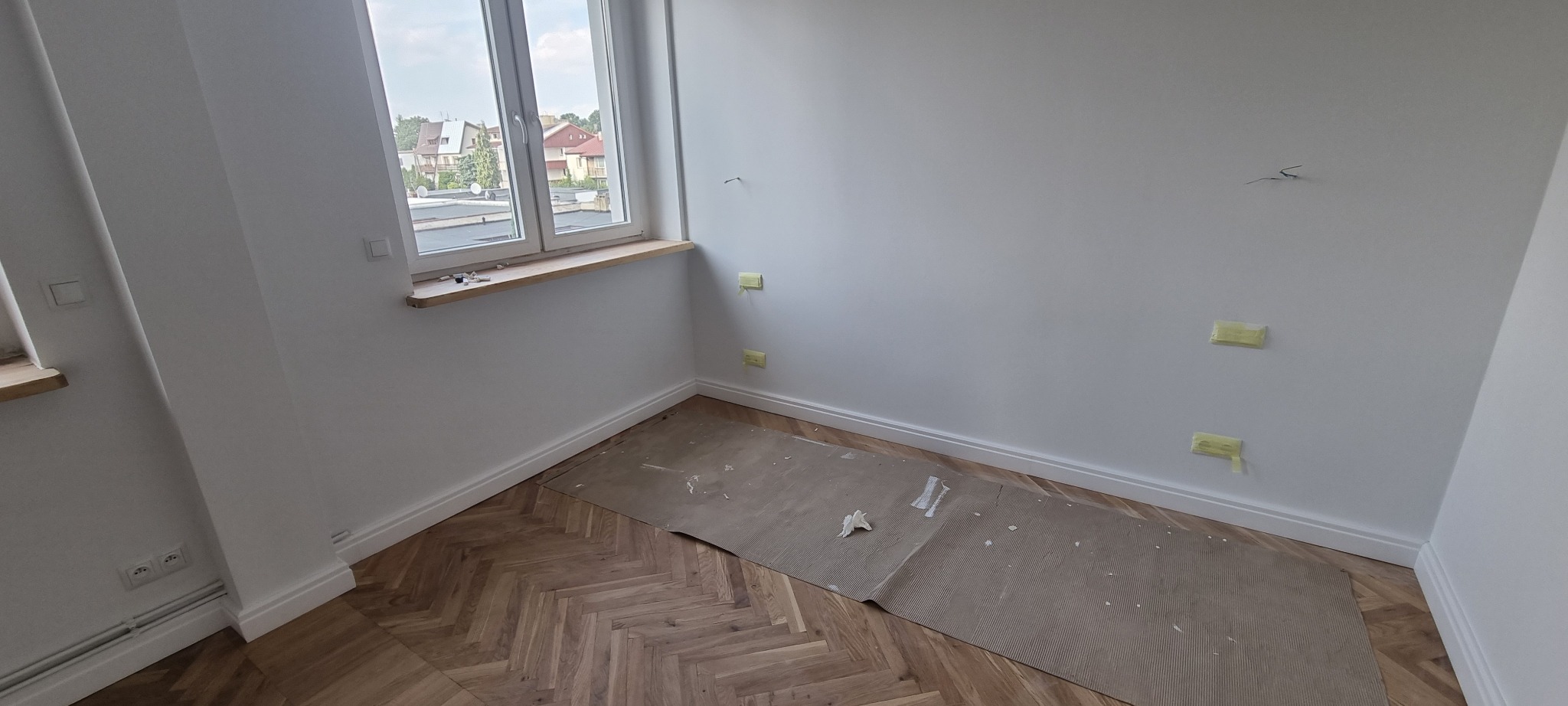 Pokój po malowaniu z zabezpieczonymi taśmą otworami na ścianie i parkietem ułożonym w jodełkę, zabezpieczonym kartonem falistym.