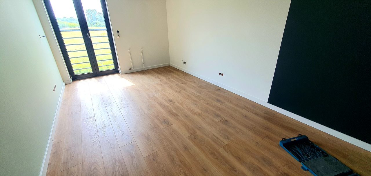 Jasne panele podłogowe ułożone w pokoju z oknem balkonowym, ściany pomalowane na biało i jasnozielono, z fragmentem ściany pomalowanym na czarno, widoczna skrzynka narzędziowa przy ścianie.