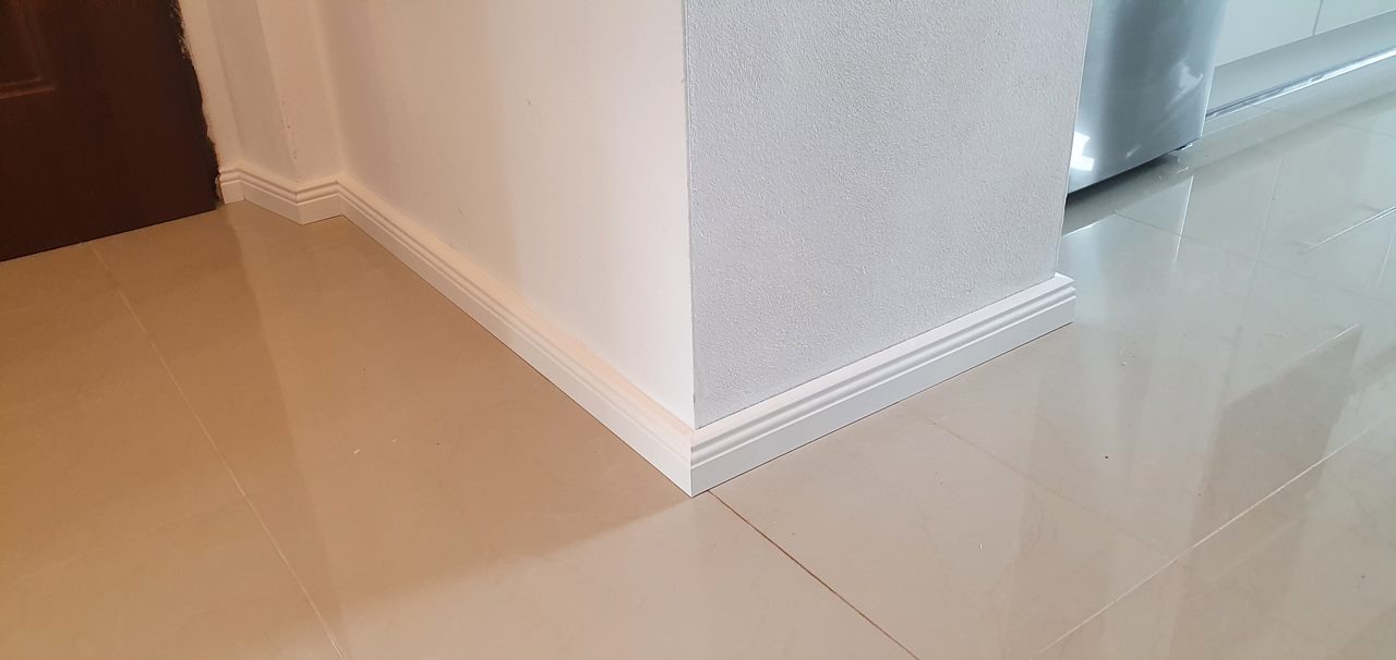 Wykończona ściana z przypodłogową listwą MDF w kolorze białym, na błyszczącej, beżowej podłodze z płytek.