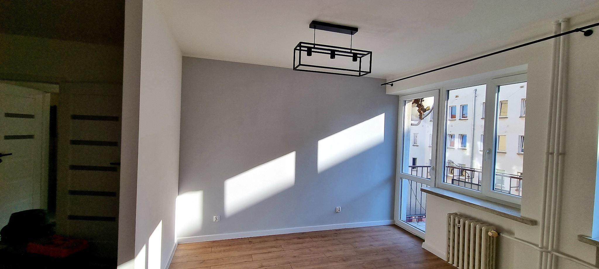 Pokój po remoncie z szarą ścianą, drewnianą podłogą, nowoczesną lampą i dużym oknem z widokiem na budynek mieszkalny.