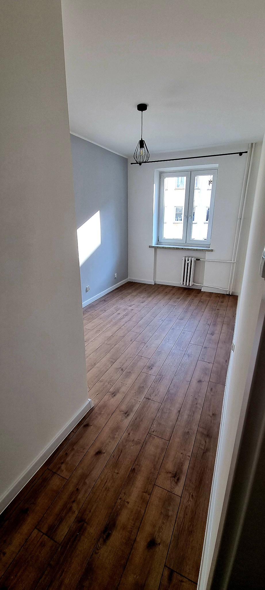 Pusta, jasna przestrzeń z nową, ciemną podłogą z paneli, szarą ścianą z białym światłem słonecznym i nowoczesną lampą wiszącą.