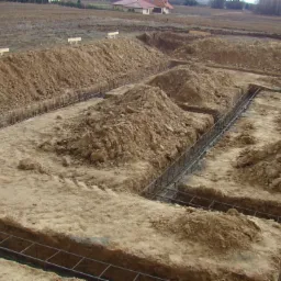 Przygotowanie terenu pod fundamenty: wykopane rowy z ułożonym zbrojeniem stalowym, ziemia usypana wzdłuż wykopów. Widok z góry na plac budowy.