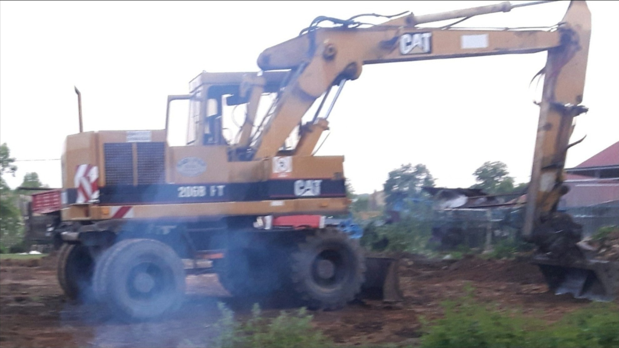 Żółta koparka kołowa CAT 206B FT z łyżką nabierającą ziemię, widoczne ślady pracy i dym, teren budowy w tle.