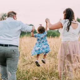 Rodzina z Mikołowa spaceruje po łące: rodzice trzymają za ręce podskakującą córeczkę w niebieskiej sukience, widok z tyłu.