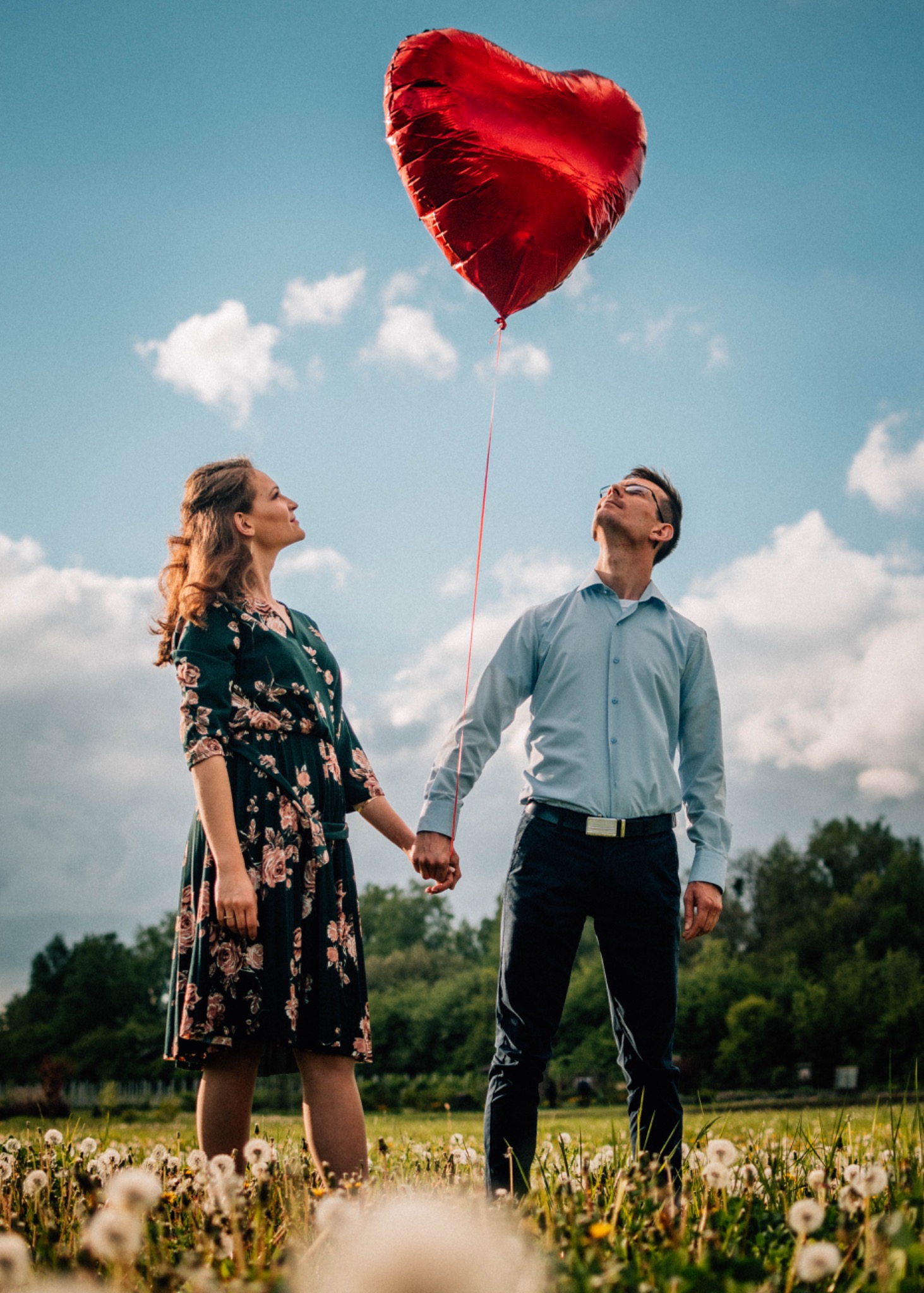 Para trzymająca się za ręce na łące dmuchawców, spoglądająca na czerwony balon w kształcie serca na tle błękitnego nieba z chmurami.