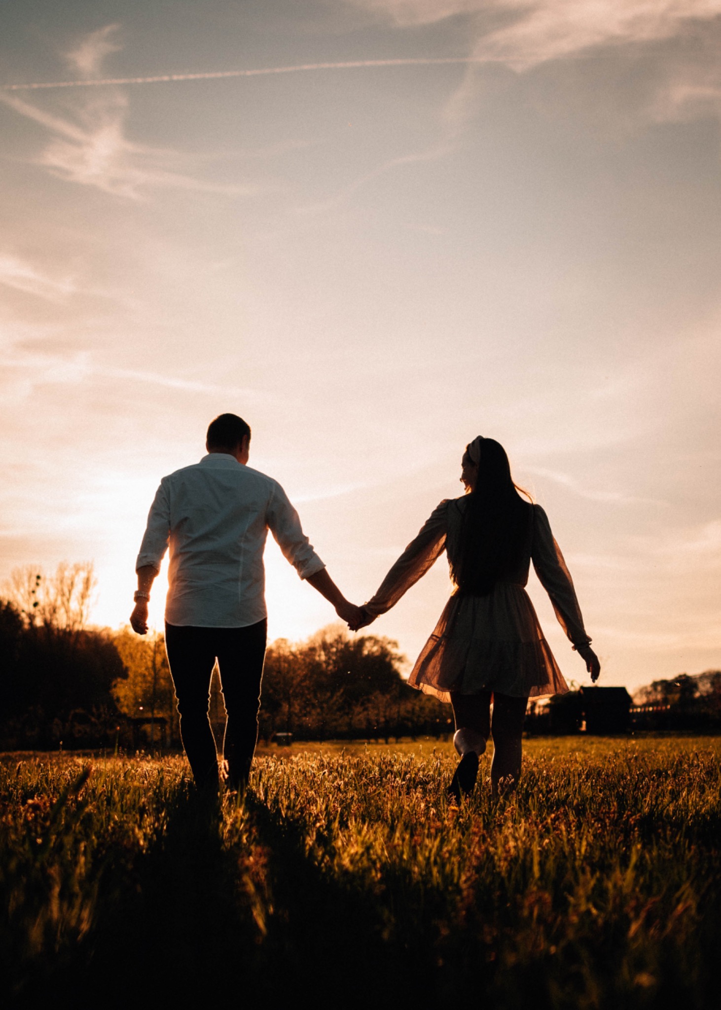 Para trzymająca się za ręce spaceruje po łące o zachodzie słońca, sylwetki na tle pomarańczowego nieba z widocznymi smugami kondensacyjnymi samolotu.