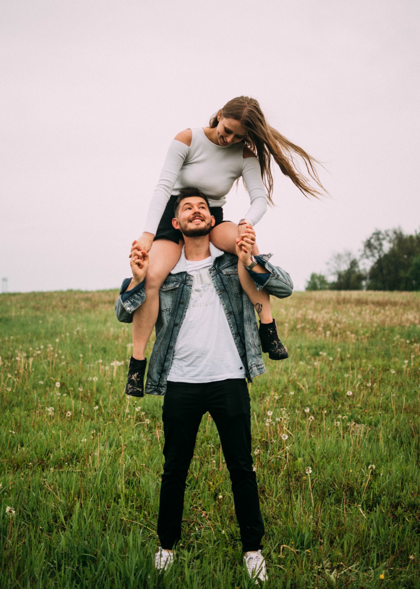 Mężczyzna z brodą w jeansowej kurtce trzyma na barana uśmiechniętą kobietę z rozwianymi włosami na łące z dmuchawcami.