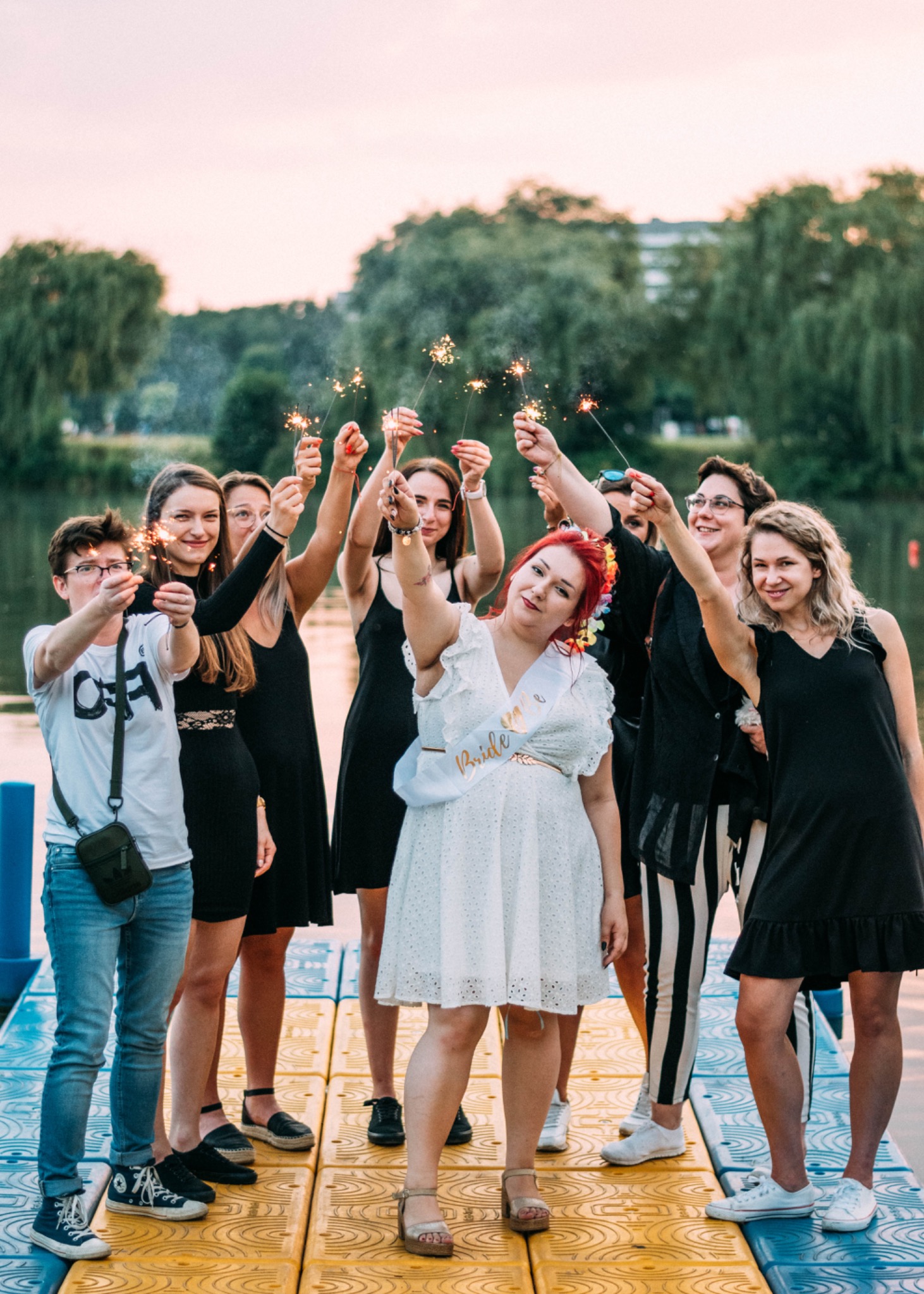 Grupa kobiet świętuje z iskierkami na pomoście nad wodą, panna młoda w białej sukience i szarfie z napisem 'Bride to be' trzyma jedną w górze, drzewa i zachodzące słońce w tle.