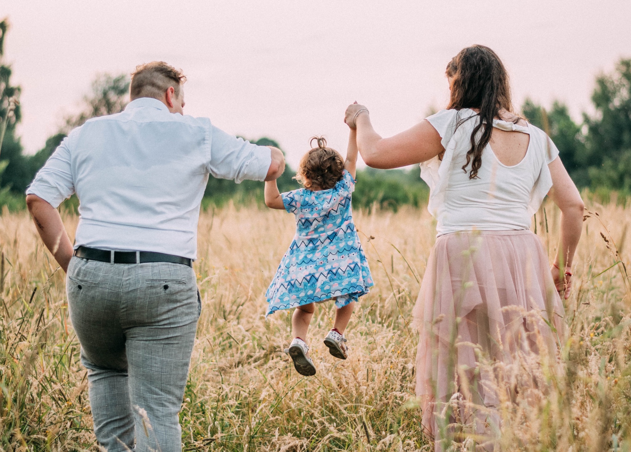 Rodzina z Mikołowa spaceruje po łące: rodzice trzymają za ręce podskakującą córeczkę w niebieskiej sukience, widok z tyłu.