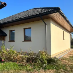 Jasna elewacja domu jednorodzinnego z ciemną dachówką, widok z boku na tle błękitnego nieba i zarośniętego ogrodu.