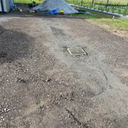 Teren po pracach ziemnych, widoczny właz kanalizacyjny, hałda kamienia oraz zabawki dziecięce w tle.