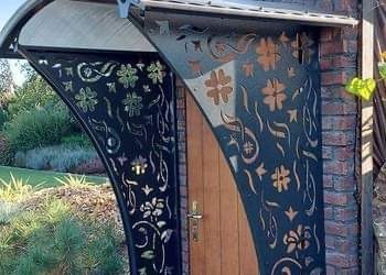 Wejście do domu z drewnianymi drzwiami osłonięte metalową konstrukcją z wycinanymi wzorami roślinnymi i geometrycznymi, zadaszone przezroczystym daszkiem, widoczne elementy muru z cegły i zieleń...