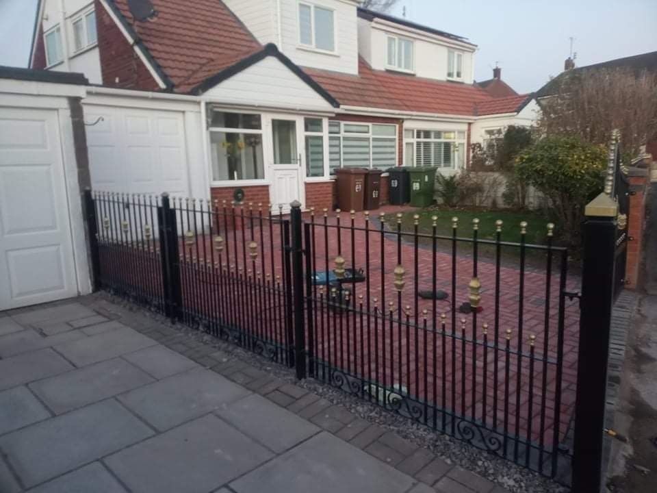 Czarna metalowa brama wjazdowa z ozdobnymi złotymi elementami na tle domu z czerwoną dachówką i brukowanym podjazdem.