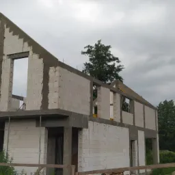 Surowy stan dwukondygnacyjnego domu z białych bloczków, widoczne otwory okienne i drzwiowe, szare niebo w tle, tablica ostrzegawcza 'Uwaga teren budowy' w prawym dolnym rogu.