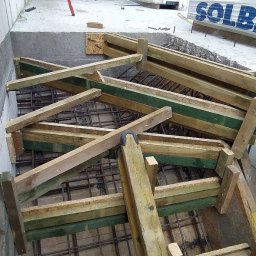 BiałoBud - Zbrojenie fundamentu z drewnianym szalunkiem, widoczne pręty zbrojeniowe i elementy drewniane, w tle bloczki silikatowe Solbet i poziomica.