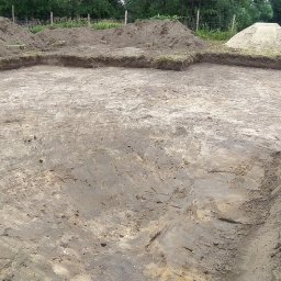 BiałoBud - Wytyczony teren pod budowę, widoczne wykopane obrysy fundamentów z zachowanym darniem, w tle pryzmy ziemi i fragment koparki gąsienicowej.