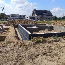 Zalane betonem fundamenty domu jednorodzinnego z hydroizolacją, widoczne wykopy pod fundamenty i wystające betonowe słupy, w tle zabudowa jednorodzinna i błękitne niebo.