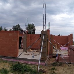 BiałoBud - Wznoszenie ścian parteru domu jednorodzinnego z czerwonej cegły Porotherm, widoczne zbrojenia słupów i podpory montażowe, w tle pracownicy na dachu.