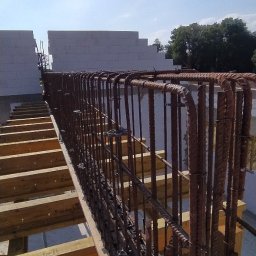 BiałoBud - Zbrojenie pionowe i poziome przygotowane do zalania betonem, widoczne szalunki drewniane i bloczki betonowe w tle, słoneczny dzień.