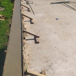 BiałoBud - Świeżo wylana, mokra wylewka betonowa w szalunku z drewnianymi podporami, widok z góry na plac budowy z zielonym tłem.