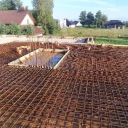 Siatka zbrojeniowa na płycie fundamentowej z widocznymi pionowymi prętami i deskowaniem, częściowo zalana wodą, w tle zabudowa jednorodzinna.