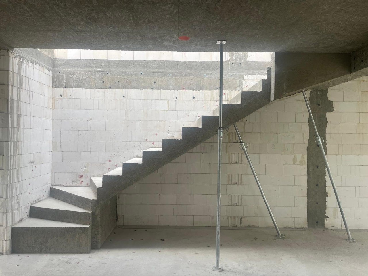 Surowe betonowe schody w trakcie budowy, wsparte metalowymi stemplami, na tle ściany z białych bloczków.