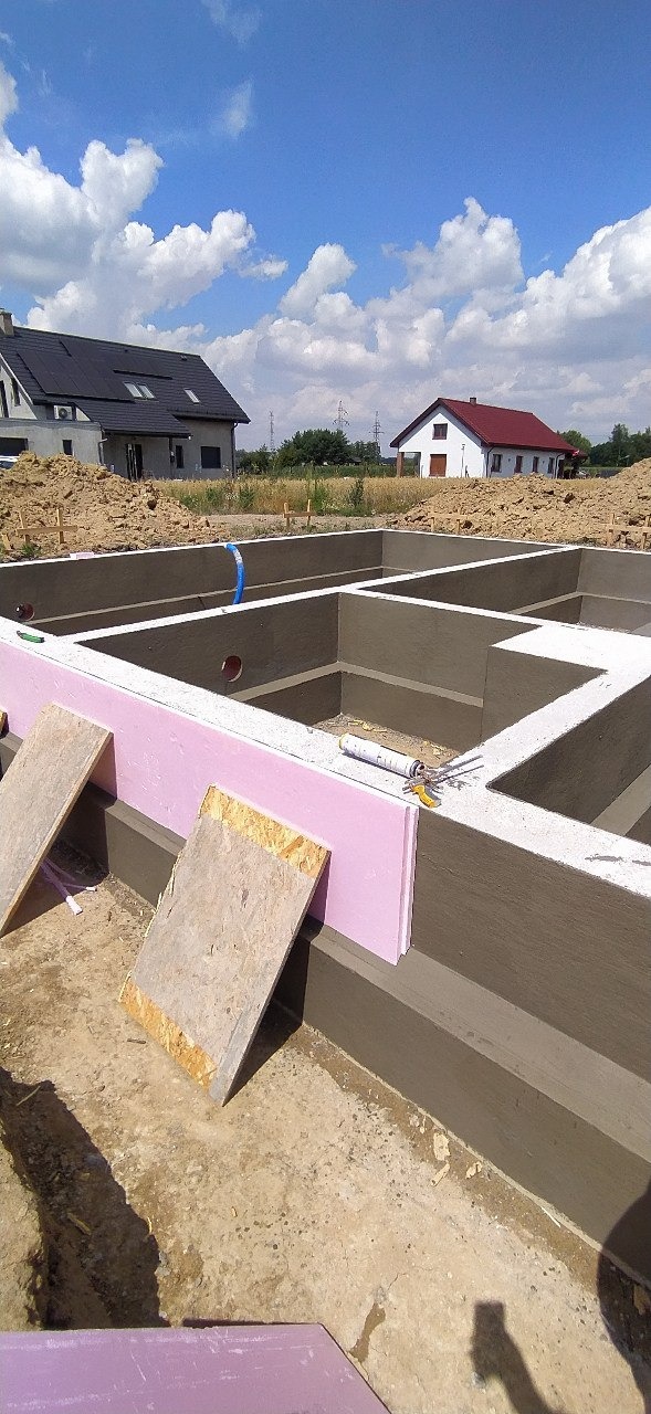 Wykop pod fundamenty domu jednorodzinnego z częściowo ułożoną izolacją termiczną w postaci różowego styropianu oraz widocznymi deskami szalunkowymi, w tle domy i błękitne niebo z chmurami.