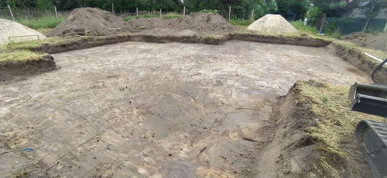 Wytyczony teren pod budowę, widoczne wykopane obrysy fundamentów z zachowanym darniem, w tle pryzmy ziemi i fragment koparki gąsienicowej.