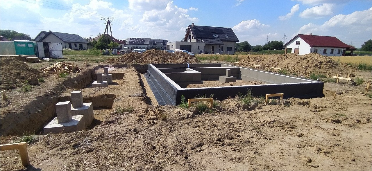 Zalane betonem fundamenty domu jednorodzinnego z hydroizolacją, widoczne wykopy pod fundamenty i wystające betonowe słupy, w tle zabudowa jednorodzinna i błękitne niebo.