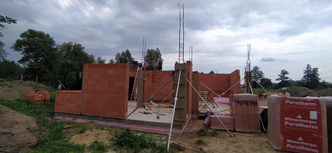 Wznoszenie ścian parteru domu jednorodzinnego z czerwonej cegły Porotherm, widoczne zbrojenia słupów i podpory montażowe, w tle pracownicy na dachu.