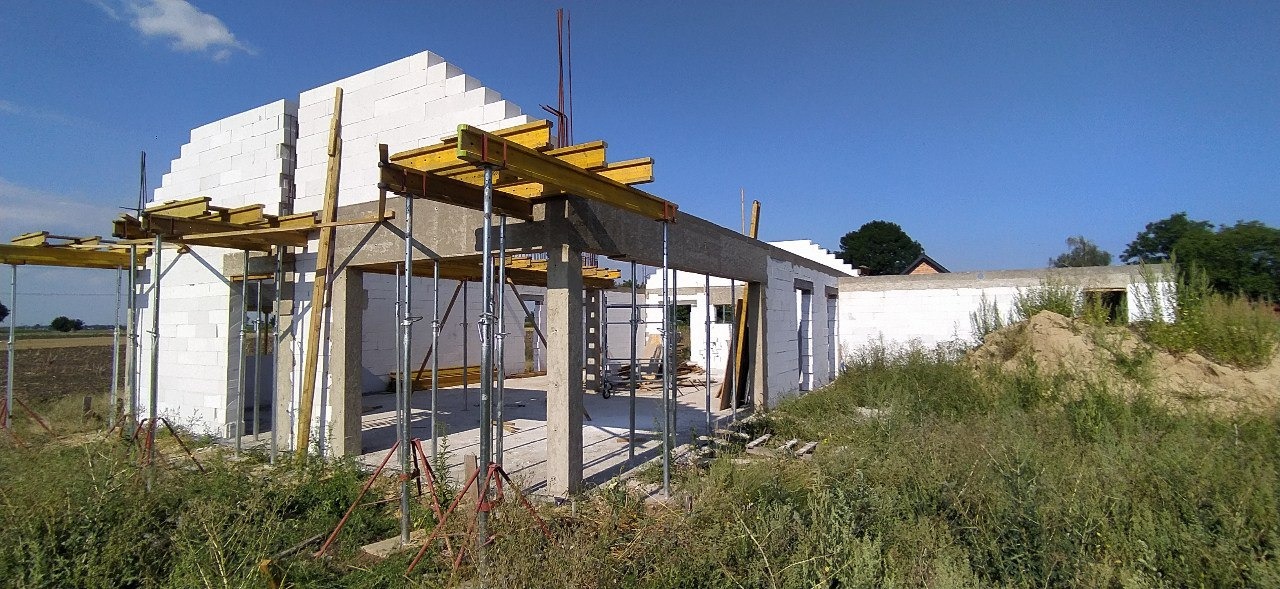 Budowa domu z białych bloczków, widoczne zbrojenia i podpory stropu, otoczenie w stanie surowym.