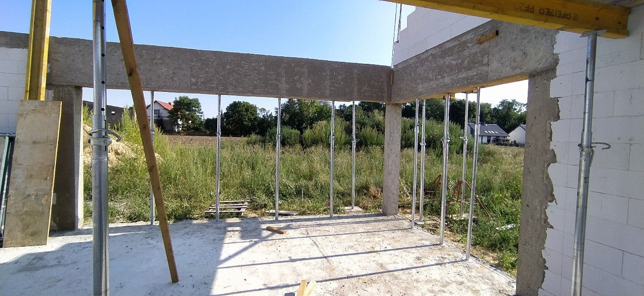 Surowy stan budynku z betonu komórkowego, widoczne zbrojenia i podpory stalowe, otoczenie zielone, domy w tle.