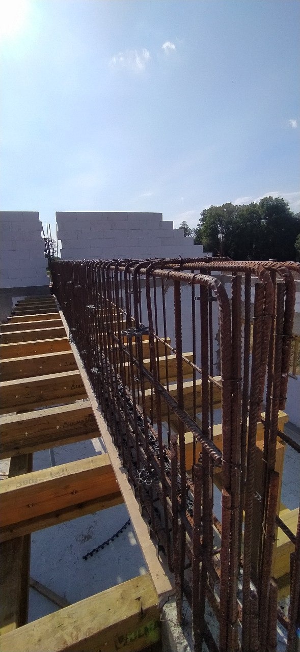 Zbrojenie pionowe i poziome przygotowane do zalania betonem, widoczne szalunki drewniane i bloczki betonowe w tle, słoneczny dzień.