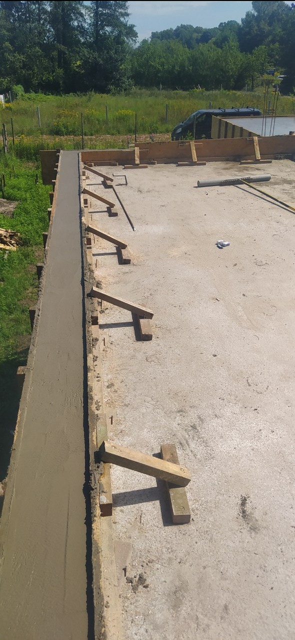 Świeżo wylana, mokra wylewka betonowa w szalunku z drewnianymi podporami, widok z góry na plac budowy z zielonym tłem.