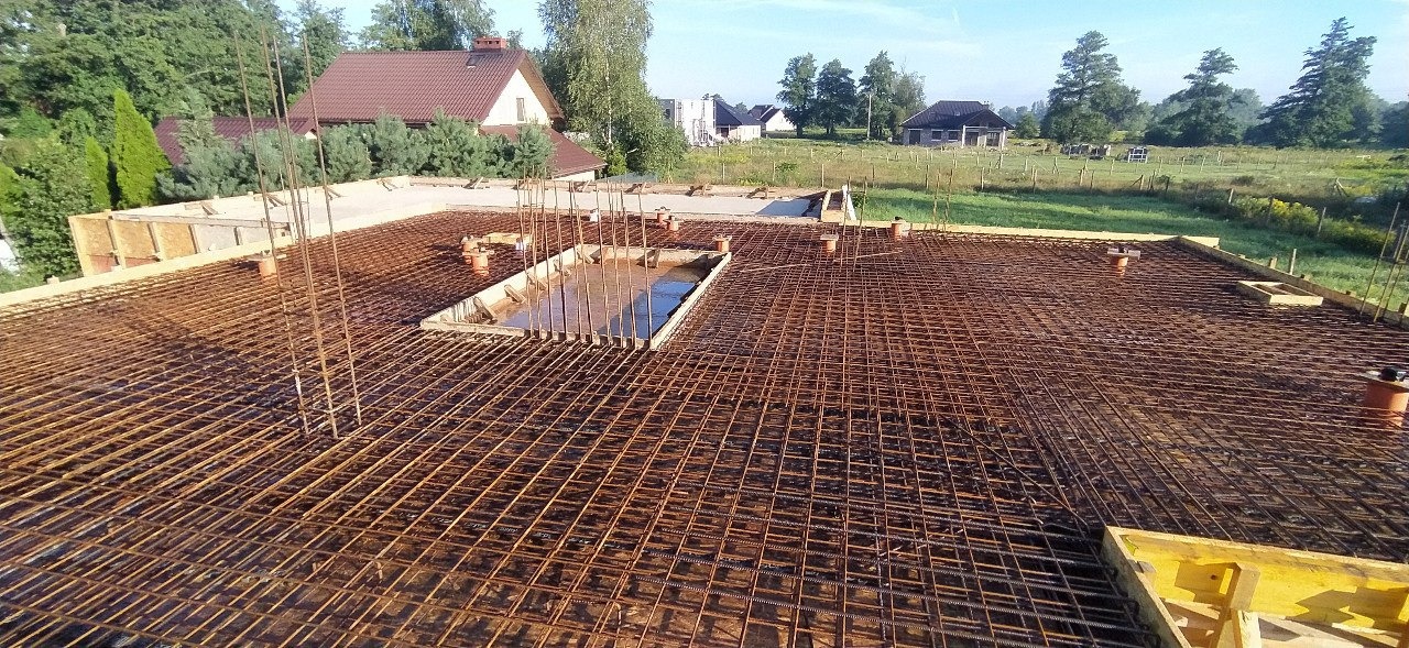 Siatka zbrojeniowa na płycie fundamentowej z widocznymi pionowymi prętami i deskowaniem, częściowo zalana wodą, w tle zabudowa jednorodzinna.