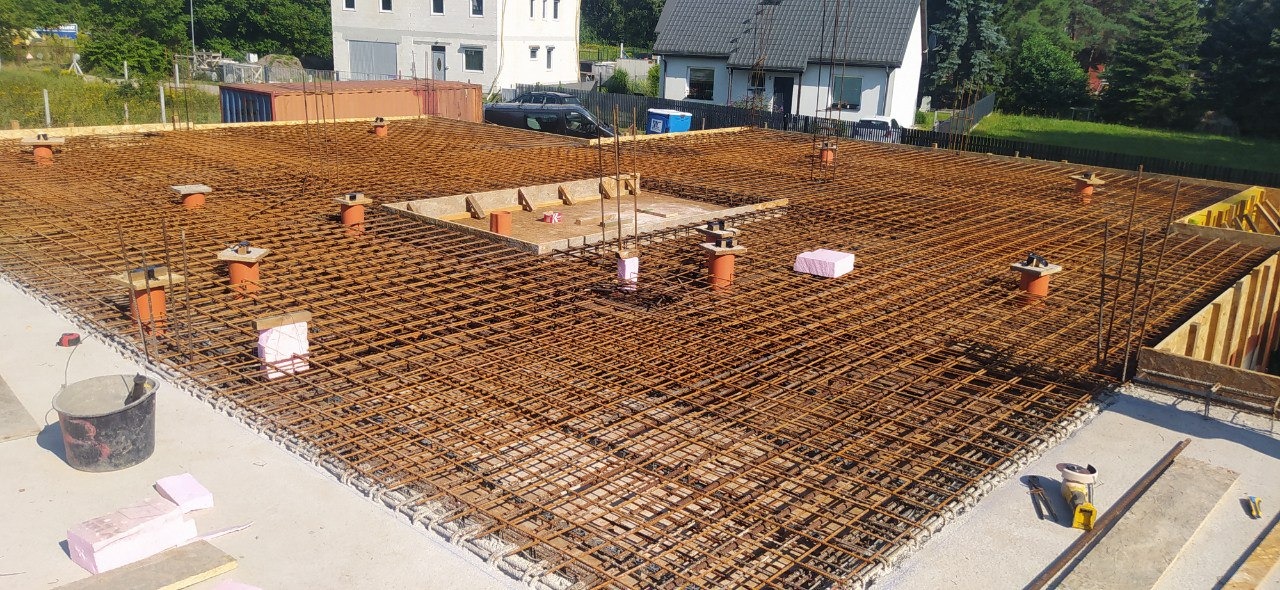 Siatka zbrojeniowa przygotowana do zalania betonem na fundamencie budynku, widoczne podpory z pomarańczowych rur, szalunki na obrzeżach i w centralnej części, oraz budynki mieszkalne w tle.