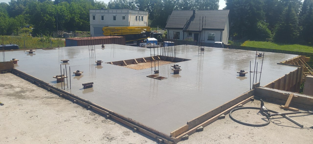 Świeżo wylany, mokry fundament z wystającymi prętami zbrojeniowymi i drewnianym szalunkiem, na tle budynku z żółtym dźwigiem budowlanym.