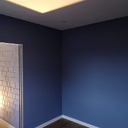 Pomieszczenie po malowaniu ścian na granatowo, zabezpieczona folią lampa sufitowa, widoczna imitacja cegieł na ścianie obok drzwi, drewniana podłoga.